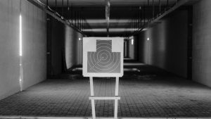 Black and white photo of a shooting range with a focus on a paper target in a dimly lit indoor setting.