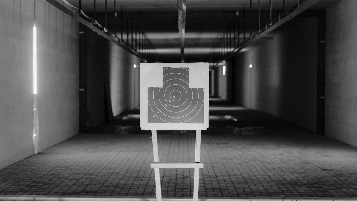 Black and white photo of a shooting range with a focus on a paper target in a dimly lit indoor setting.